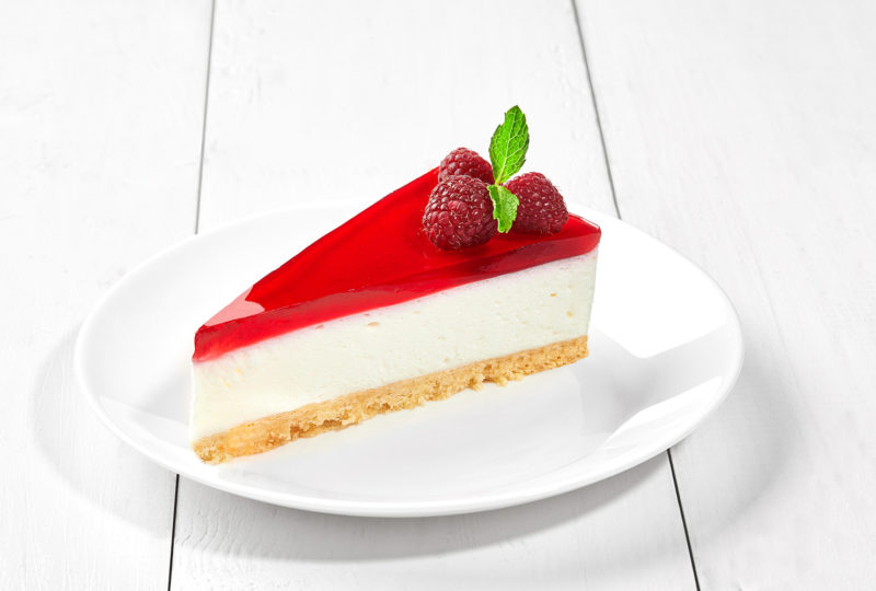 Slice of delicious delicate Philadelphia cheesecake on shortcrust pastry with raspberry jelly garnished with fresh berries and fragrant mint leaves served on plate on white wooden background